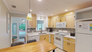 Une cuisine moderne avec des meubles blancs et jaunes, un évier en acier inoxydable, un four à micro-ondes, une cuisinière et un réfrigérateur. On y trouve une table à manger avec des chaises et un luminaire suspendu.
