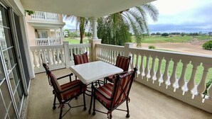 Varanda coberta com mesa e cadeiras, balaústres brancos e vista para uma paisagem verde.