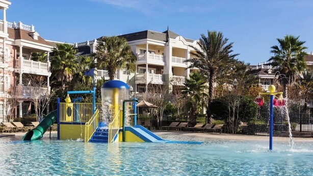 Piscina com escorrega e área de recreação aquática, cercada por palmeiras e edifícios de hotel.