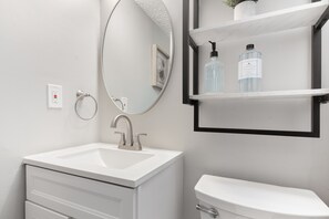 Une salle de bain avec un lavabo blanc, un miroir rond, des toilettes et une étagère contenant des articles de toilette.