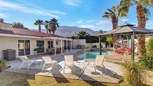 Un patio trasero con piscina, sillas de descanso, un área de estar cubierta y palmeras.