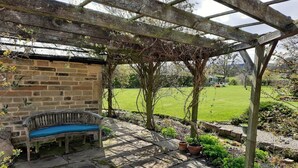 A stone bench under a wooden pergola with a brick wall backdrop, a grassy yard, and a clear sky.