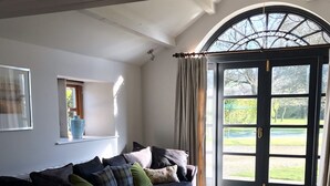 A cozy living room with a dark blue sofa, a wooden coffee table, and a dining table. A large window with curtains provides a view of a garden.
