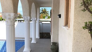 A covered walkway with white columns, a pool, and a view of palm trees. - Modern 4-bedroom villa, private pool, near beach.  (Empuriabrava)