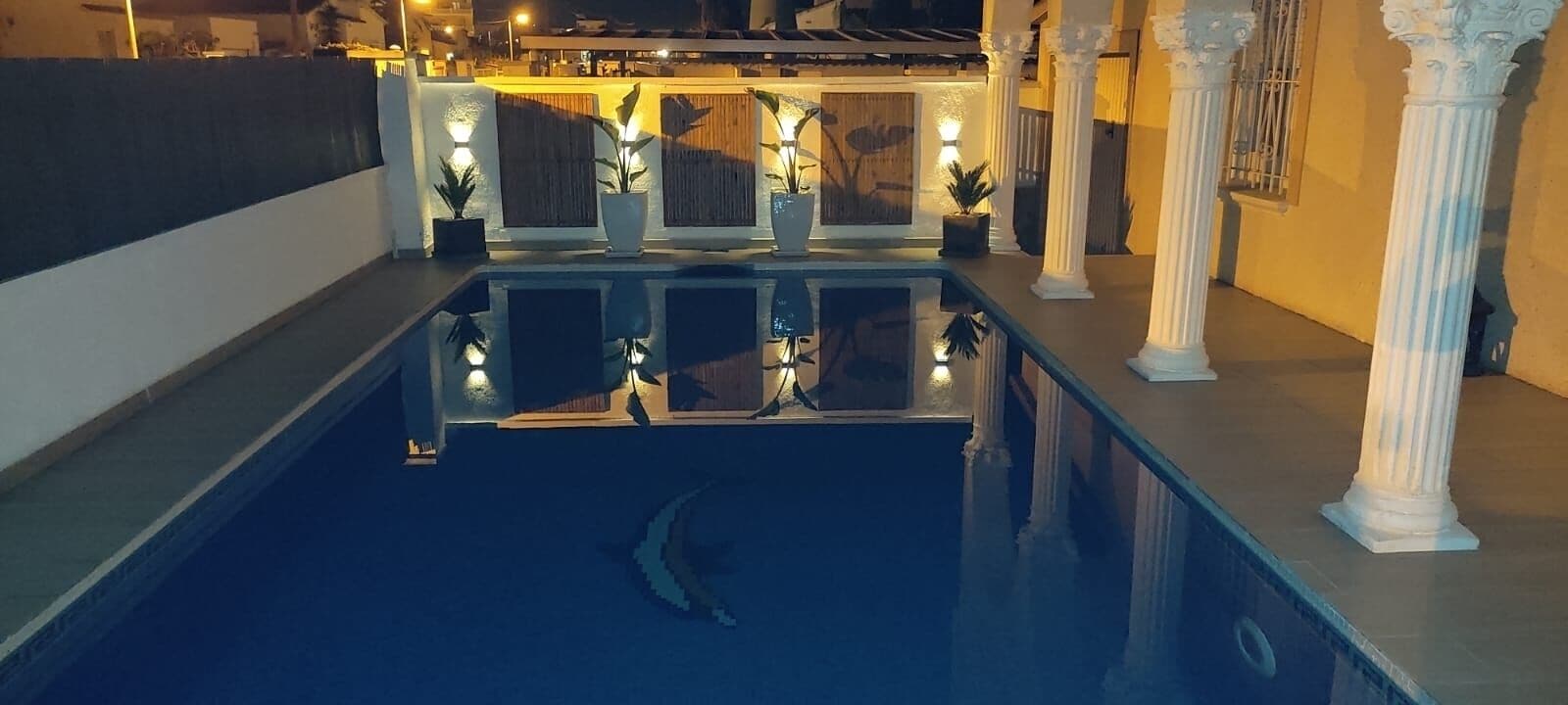 Un área de piscina con una escultura de delfín, columnas blancas y plantas en macetas.