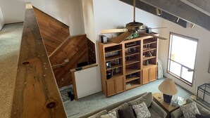 A spacious living area with a wooden staircase, a built-in bookshelf, a ceiling fan, and a large window providing natural light. - SM BBR home - Perfect for two families! (Sisters)