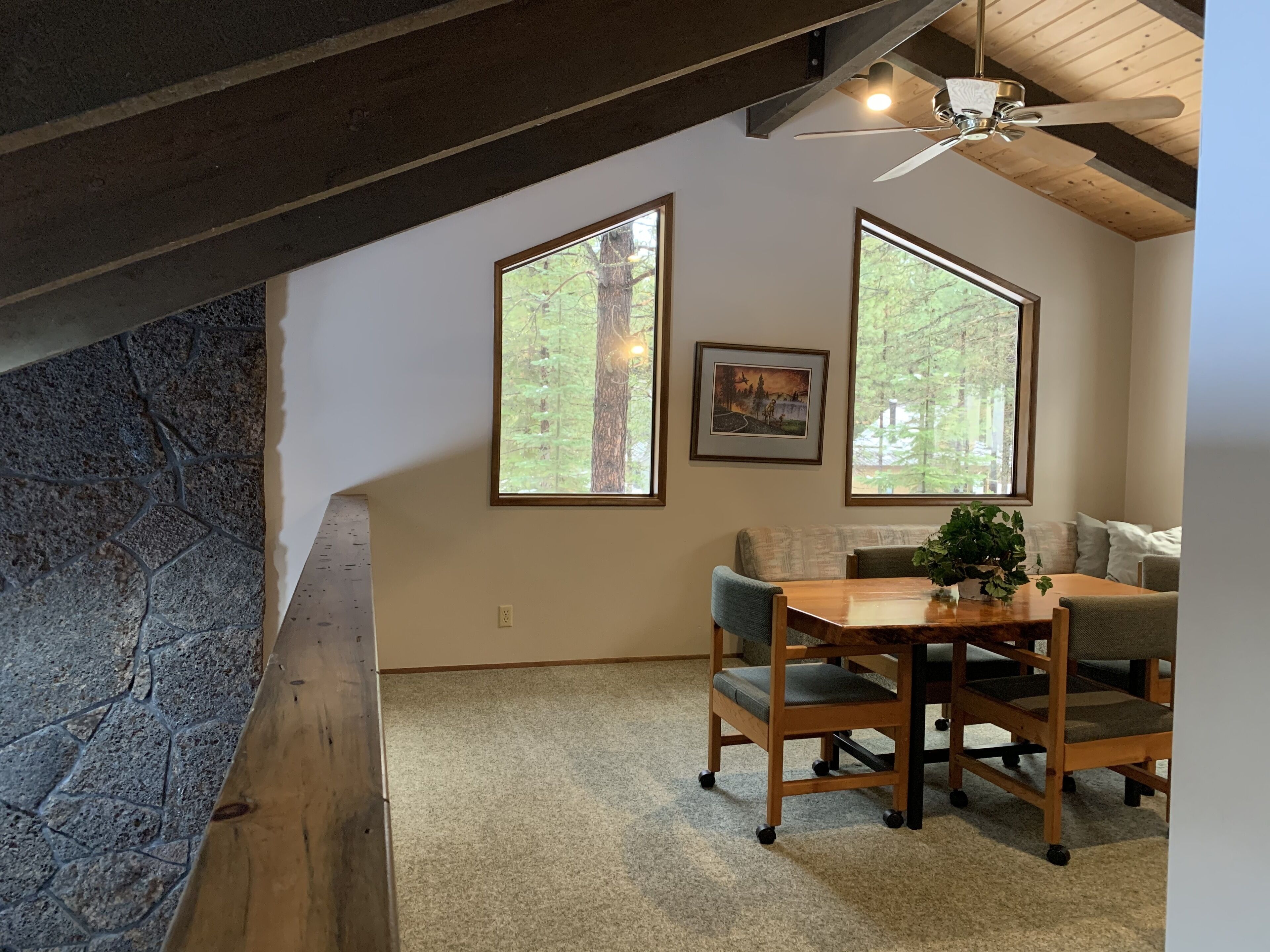 A cozy room with a stone wall, a wooden table with chairs, a ceiling fan, and large windows with a forest view.