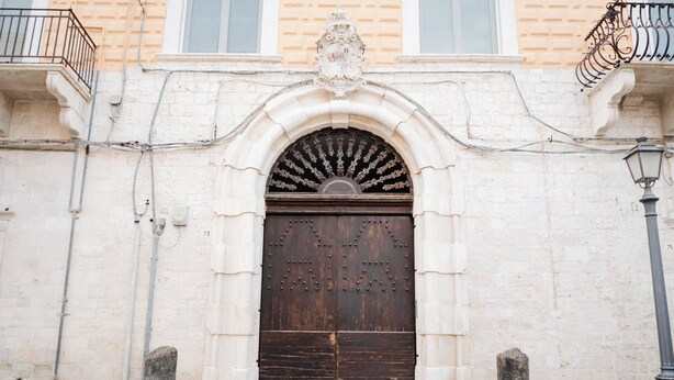 Un edificio histórico con una puerta de madera grande y ornamentada, fachada de piedra y un balcón con barandilla de hierro forjado.