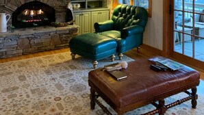 Una sala de estar acogedora con una chimenea de piedra, un sillón verde, un reposapiés de cuero y un televisor sobre la chimenea.