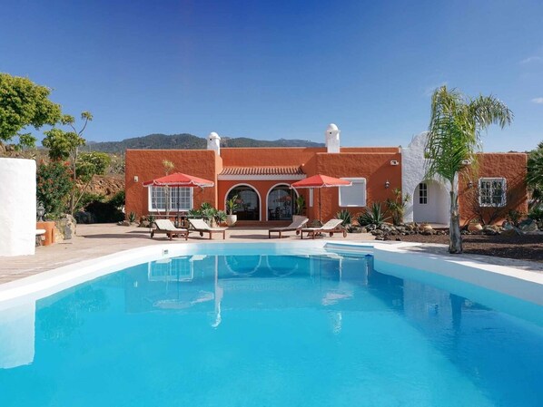 Vue sur une villa de style méditerranéen avec piscine, transats et, en arrière-plan, des montagnes, le tout vu depuis le bord de la piscine.