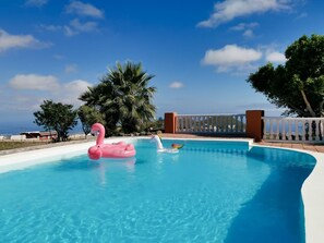 Une piscine agrémentée d’un flotteur en forme de flamant rose et d’un autre en forme d’arc-en-ciel, entourée de palmiers et d’une clôture blanche.