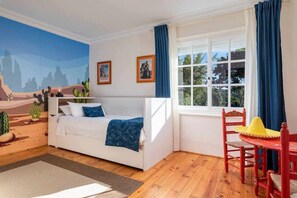 Une chambre avec un lit blanc, une fresque murale sur le thème du désert, une fenêtre avec des rideaux bleus et une chaise rouge surmontée d’un chapeau jaune.