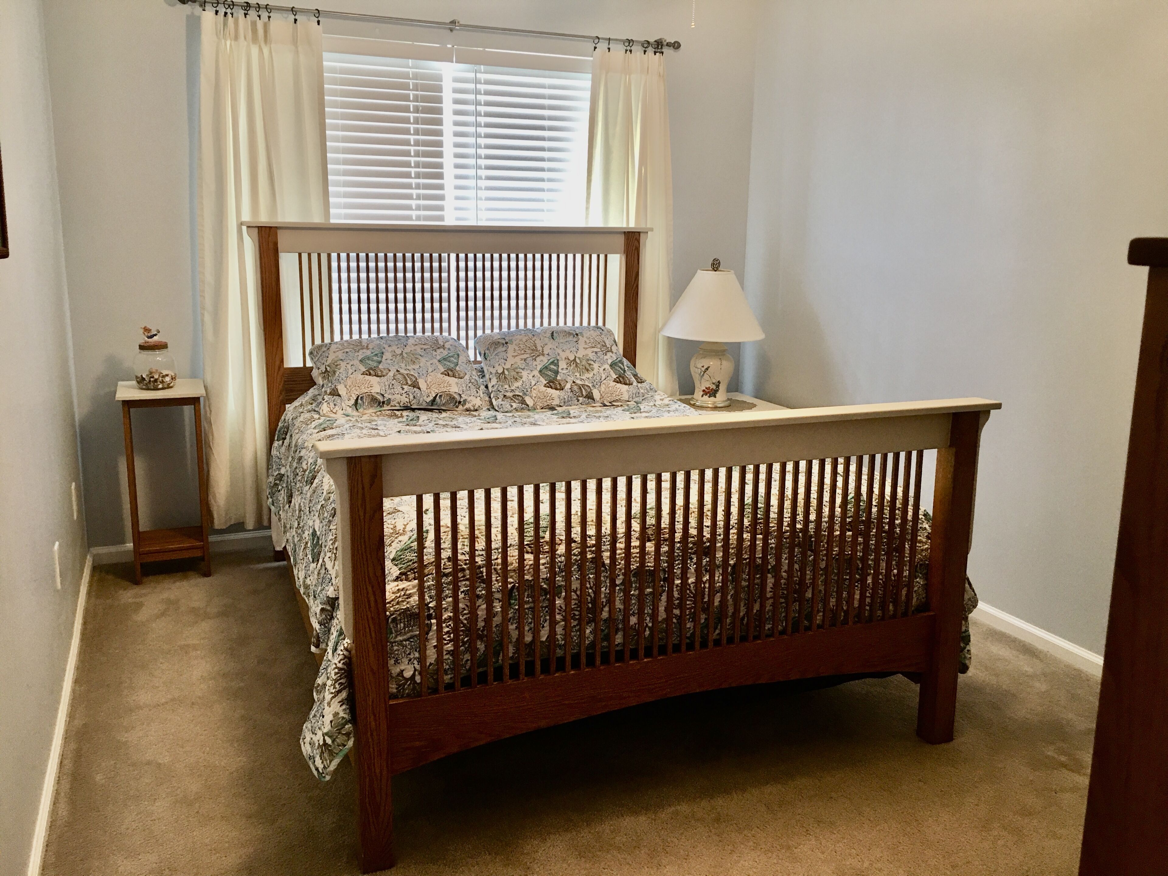 A bedroom with a wooden bed, a bedside lamp, a nightstand, and a window with blinds.