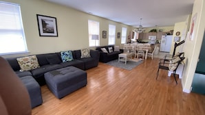 A living room with a dark-colored sectional sofa, a dining table with chairs, and a kitchen area with a refrigerator and microwave. - NEW! Corner Townhouse: 3BR, 2 balcony home, 2 blocks to beach! (Wildwood)