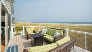 A deck with wicker furniture and a table, overlooking a beach and grassy dunes.