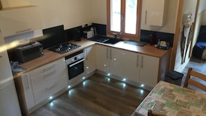 Cocina compacta con armarios blancos, microondas, estufa y pileta. Hay una ventana sobre la pileta y una mesa con mantel.
