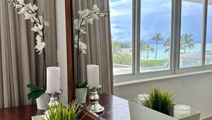 Quarto com mesa de madeira, dois vasos brancos com plantas verdes, um espelho grande refletindo palmeiras e o oceano, e duas velas brancas sobre suportes de prata.