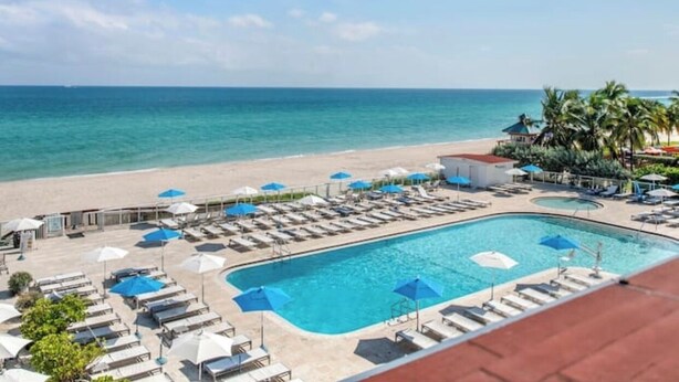 Uma área à beira da piscina com espreguiçadeiras e guarda-sóis, piscina e vista para a praia.