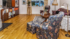 Un soggiorno con pavimento in legno, TV, libreria, una finestra con vista, una poltrona fantasia, un tavolino da caffè e una lampada.