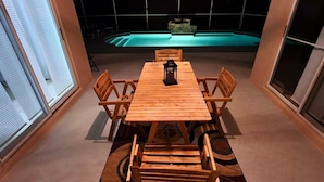 Ett matbord i trä med stolar i ett rum med stora fönster, en pool i bakgrunden och en lykta på bordet.