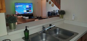 Et moderne køkken med en dobbeltvask, et fjernsyn der viser streamingtjenester, og et spiseområde, som kan ses igennem vinduet.
