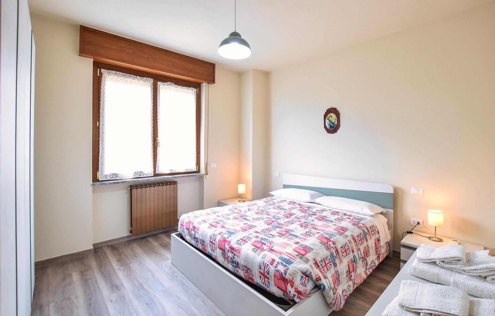 Une chambre à coucher avec un lit, une fenêtre ornée de rideaux en dentelle, un radiateur, une lampe de chevet, une horloge murale et un porte-serviettes.