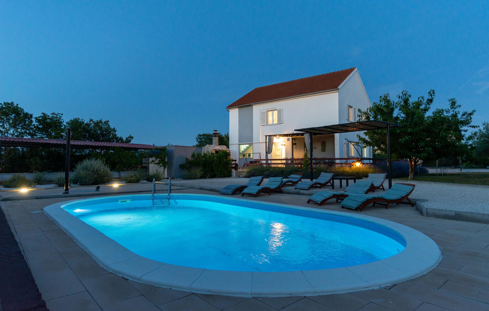 Piscina com espreguiçadeiras, um alpendre coberto e uma casa ao fundo.