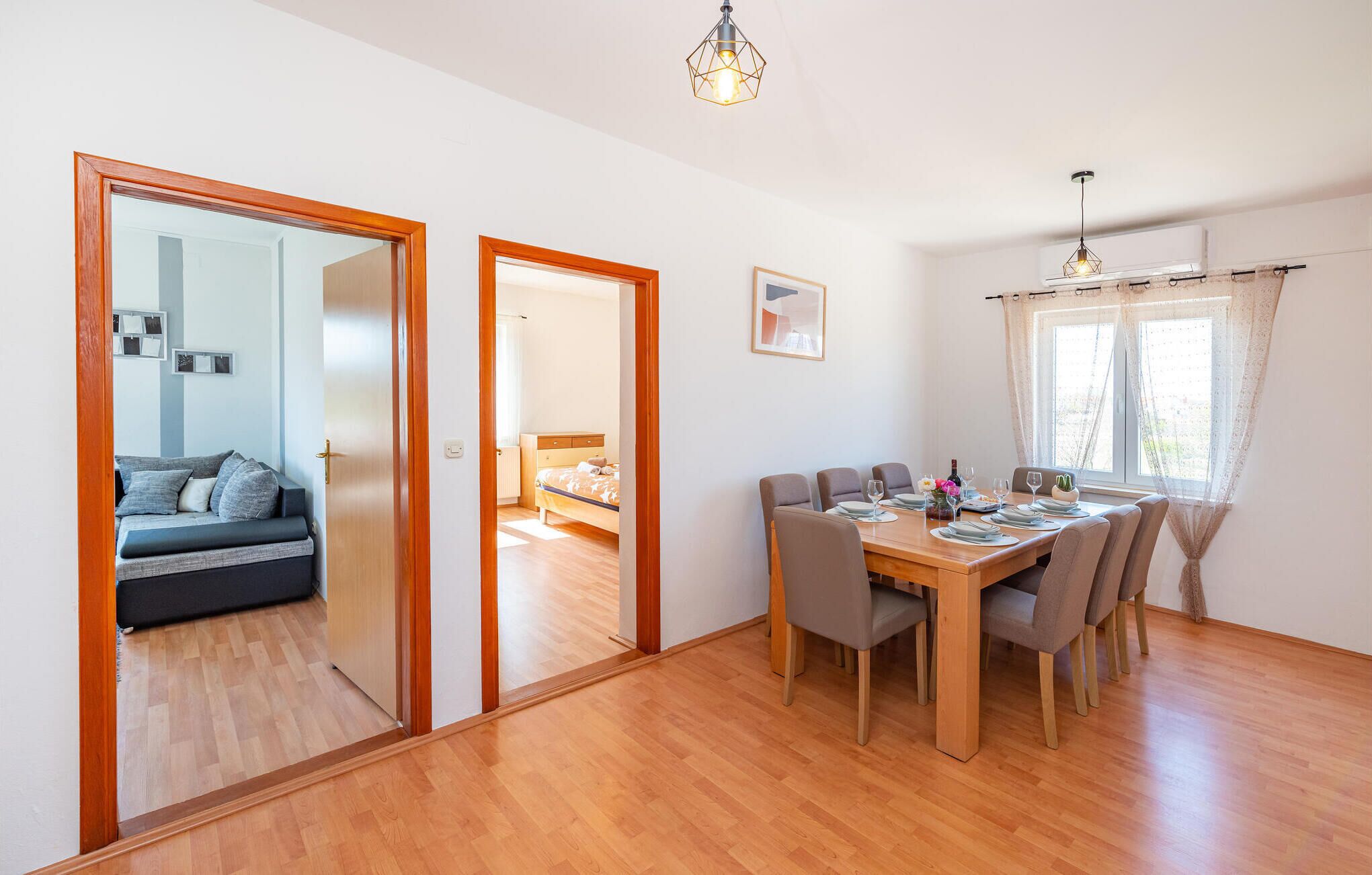 Een eetgedeelte met een houten tafel en stoelen, een bank in de aangrenzende kamer, en een hanglamp.