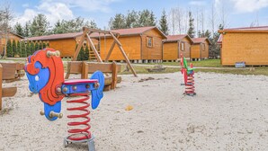 Ein Spielplatz mit zwei Schaukeln und einer Rutsche in einer Sandfläche, im Hintergrund hölzerne Hütten.