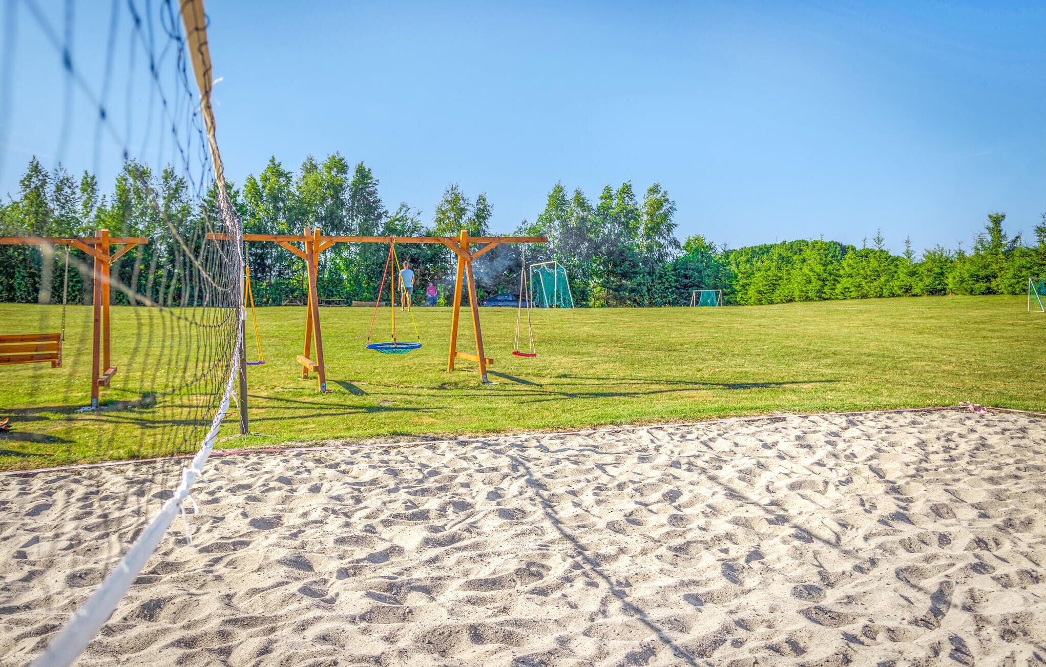 Camp de voleibol de sorra amb una xarxa, balançam nets de fusta i una porteria de futbol a un parc.