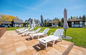 Eine Terrasse mit weißen Liegestühlen und Sonnenschirmen, ein Pool und ein gepflegter Rasen.