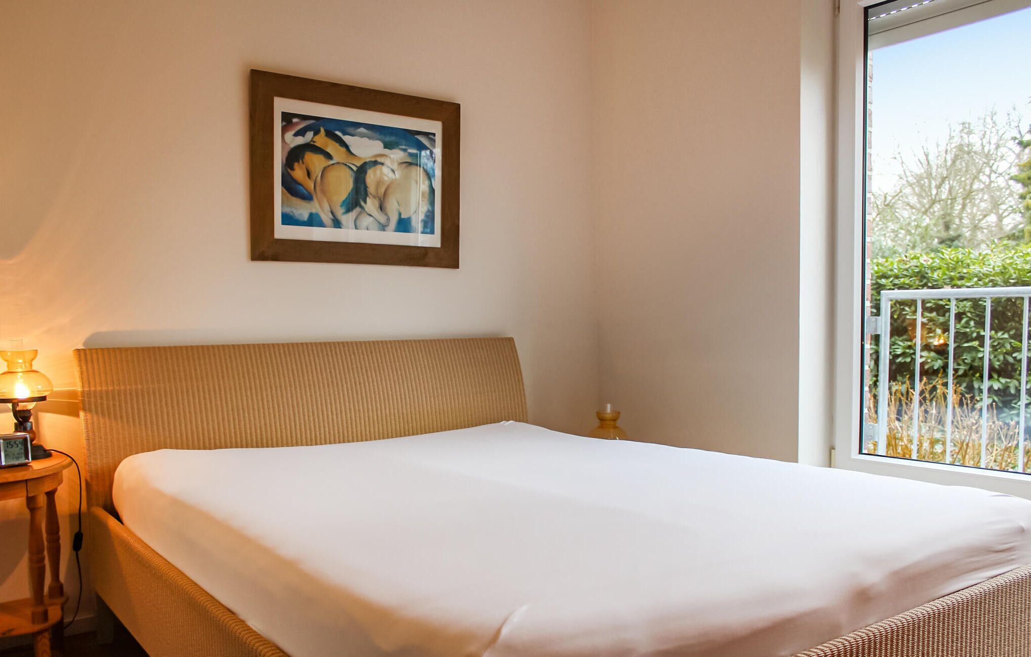 Een hotelkamer met een bed, een nachtkastje met lamp, een schilderij in een lijst aan de muur, en een balkon met beplanting.