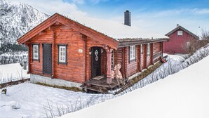Puinen mökki, jossa on musta ovi ja ikkunat, savupiippu ja taustalla punainen rakennus.