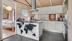 Una cocina moderna con armarios blancos, una isla central con un mapa mundial, una campana extractora de acero inoxidable y un ventanal con vistas a un área boscosa.