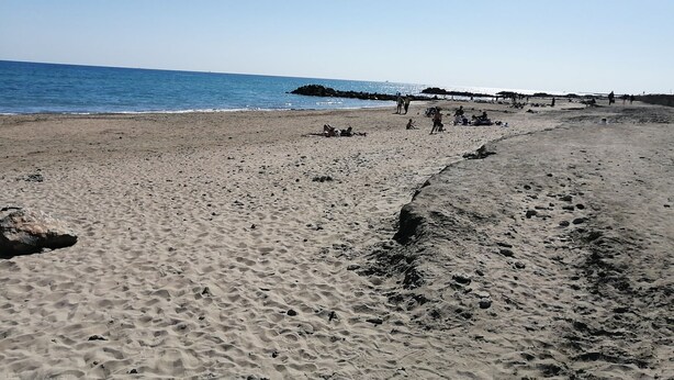 A beach with a rocky shoreline, people walking, and a pier in the background. - Maisonnette Frond de mer (Frontignan)