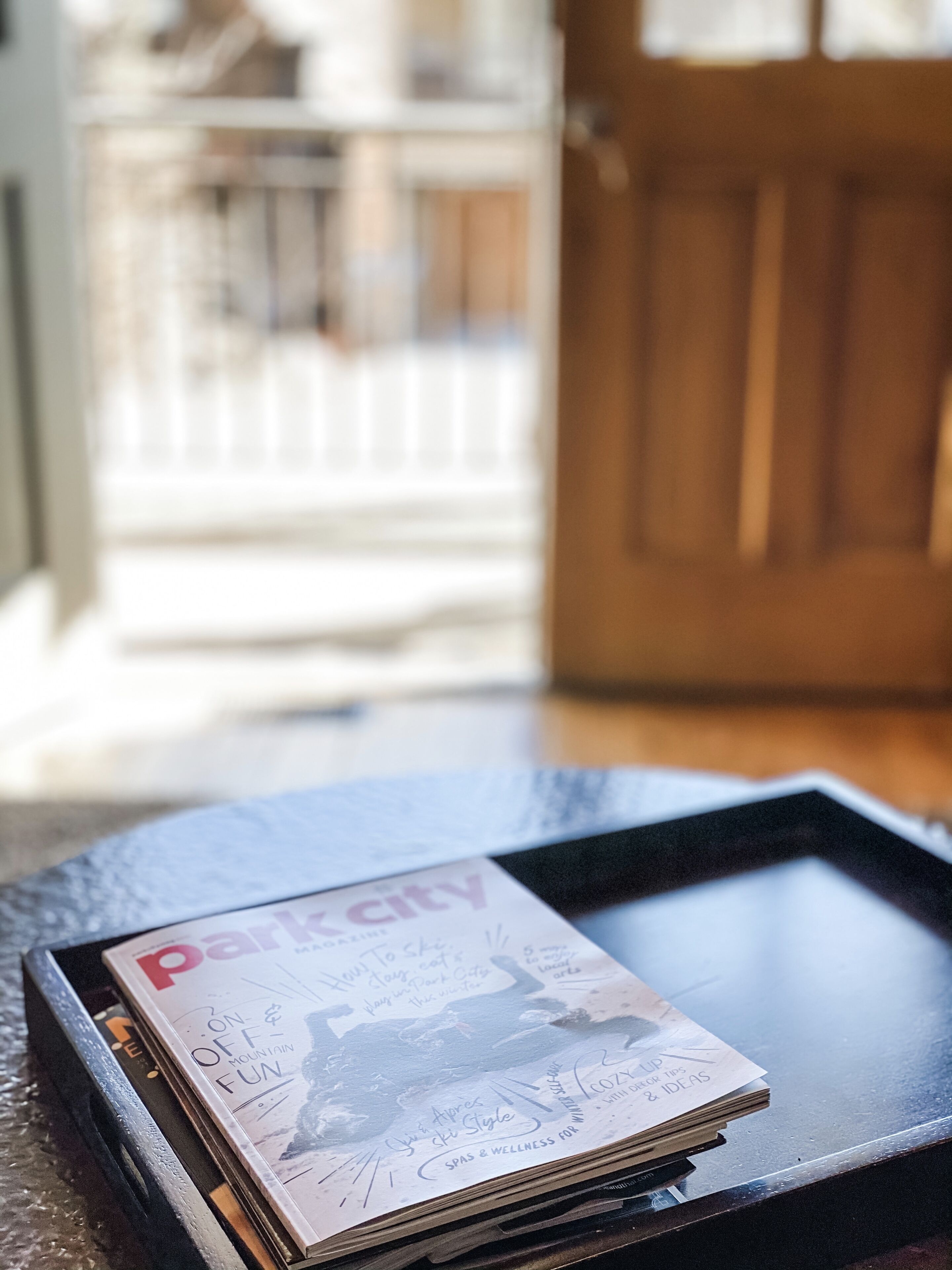 A stack of magazines on a glass table with a wooden door and railing in the background.