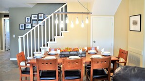 Un comedor con una mesa de madera dispuesta para seis personas, rodeada de sillas con cojines negros. Se ve una escalera con barandilla blanca al fondo.