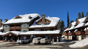 Una cabaña nevada con varias construcciones, un estacionamiento con vehículos y un cielo azul despejado.