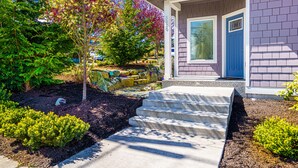 Una casa con puerta azul, un camino de piedra que lleva al frente y un jardín bien cuidado con diversas plantas y un árbol de hojas coloridas.