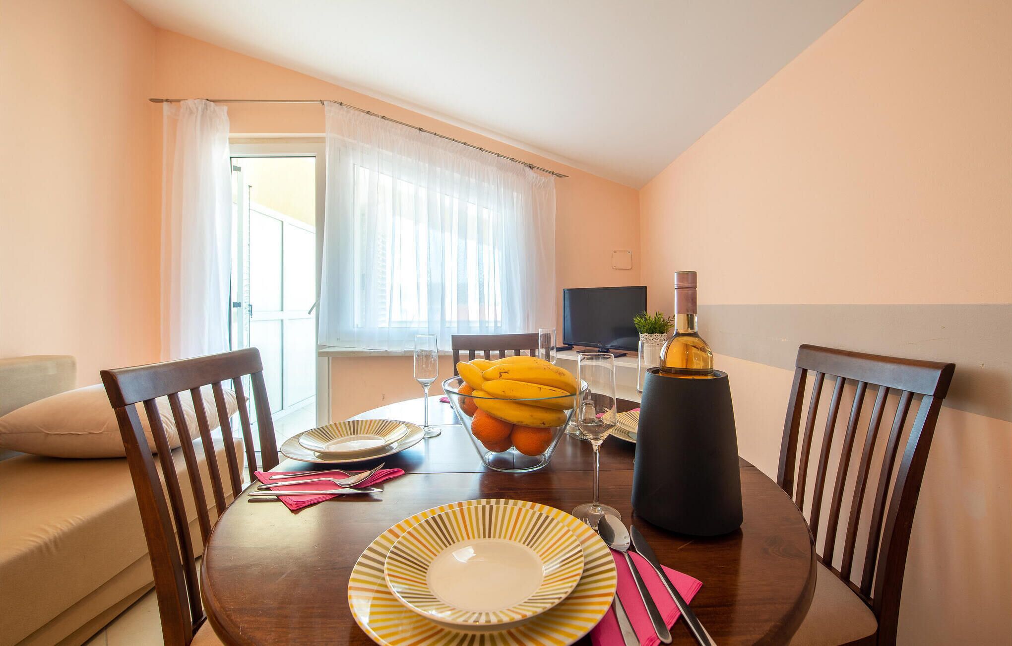 Uma área de refeições com uma mesa posta para dois, com uma tigela de frutas, uma garrafa de vinho e uma televisão ao fundo.