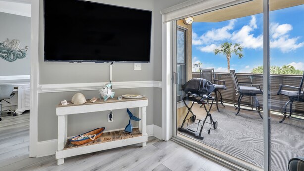 A living room with a flat-screen TV, a console table with decorative items, and a sliding glass door leading to an outdoor patio with a grill and chairs. - The SandPiper  - Ocean Front Condo (Tybee Island)