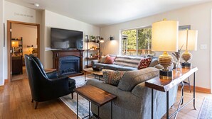 A modern living room with a fireplace, a sofa, a black armchair, and a TV. There are lamps on a side table and a window with a view of trees. - Clear Creek Mountain Cabin With Lake Access (Polson)