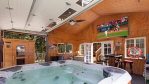 A hot tub in a wooden cabin with a television displaying a baseball game, a bar area, and a staircase leading to an upper level. - Clear Creek Mountain Cabin With Lake Access (Polson)