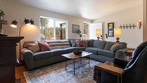 A modern living room with a sectional sofa, a coffee table, and a fireplace. There are two windows providing natural light and a view of the outdoors. - Clear Creek Mountain Cabin With Lake Access (Polson)