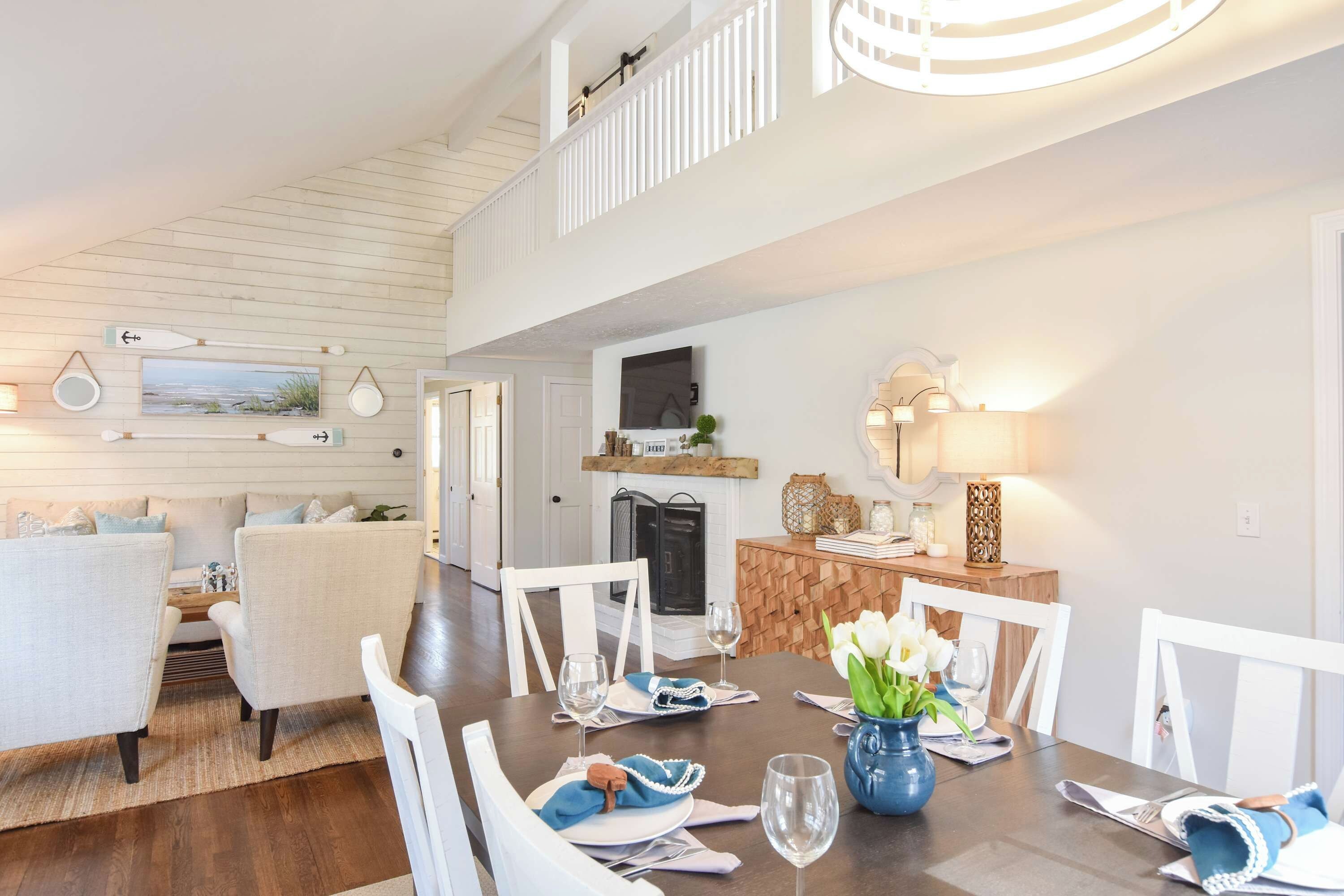 A dining area with a wooden table set for four, white chairs, a fireplace, and a television mounted above the fireplace.