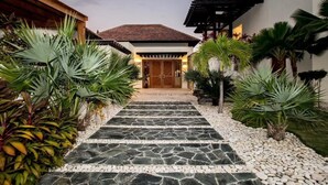 Un chemin menant à une maison avec un toit en tuiles et des portes en bois, entourée de plantes tropicales et bordée d’un sentier en pierre.