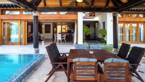 Une terrasse extérieure couverte avec une piscine, des meubles en bois et une vue sur l’intérieur.