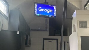 Un pasillo con un televisor montado mostrando el logotipo de Google, una puerta con un letrero de 'No Fumar' y un refrigerador.