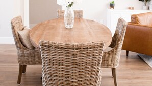 A round wooden dining table with wicker chairs, a vase with flowers, and a white wall with a picture frame and shelves with plants.