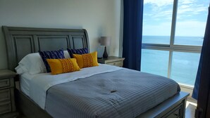 A bed with a grey headboard, white and yellow pillows, and a grey quilt. There is a nightstand with a lamp and a view of the ocean through a window with blue curtains. - OCEAN VIEW APARTMENT on the beach (Nueva Gorgona)